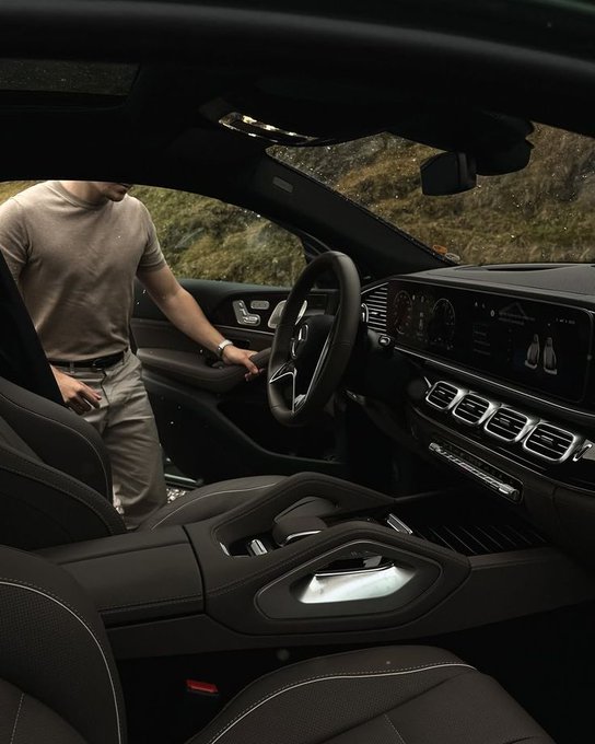 A man in a beige t-shirt stands beside a luxury Mercedes-Benz SUV, reaching toward the steering wheel in a moody, cinematic shot of the vehicle's sleek black interior featuring a dual-screen digital dashboard. The image complements the "you'll be so rich" message by evoking aspirations of wealth and success, with the high-end car symbolizing the lifestyle milestone being wished upon the recipient.