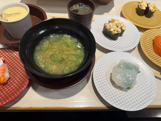 A Japanese meal featuring a bowl of miso soup with green onions, various sushi rolls on small plates, chawanmushi, and what appears to be a translucent sushi piece.