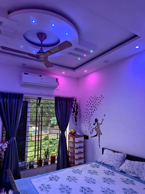 A bedroom interior features a decorative ceiling with recessed lighting and a fan, illuminated by purple and pink ambient lights, with a window, dark curtains, and wall decals visible.