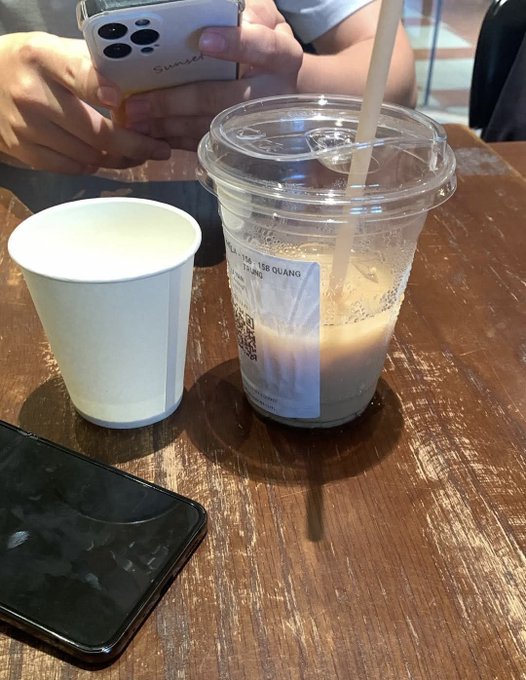 A casual photo at a café table showing a clear plastic iced coffee cup from 'TSE QUANG' next to a white paper cup, with a person holding an iPhone in the background.