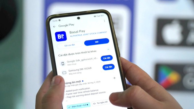 A close-up of a hand holding a smartphone showing a Google Play store page for 'Basil Pay' by ALPHARISE JOINT STOCK COMPANY, with installation options and a Vietnamese interface.