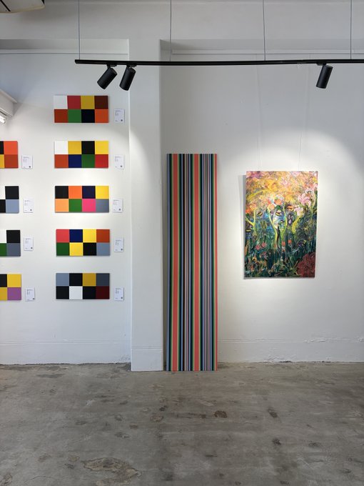 An art gallery displays several pieces, including a wall of color block paintings, a tall vertical piece with colorful stripes, and a framed impressionist-style floral painting illuminated by track lighting.