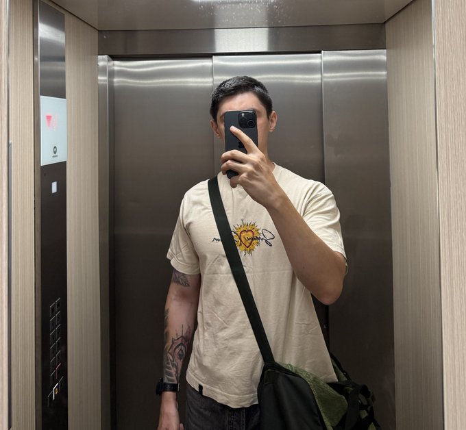 A person takes a mirror selfie inside an elevator, wearing a cream-colored t-shirt with a "Coggin's" graphic and carrying a dark messenger bag slung across their body. The image relates to the post about the bag feeling heavier, showing the bag clearly weighed down on their shoulder in a typical elevator moment to start the day.