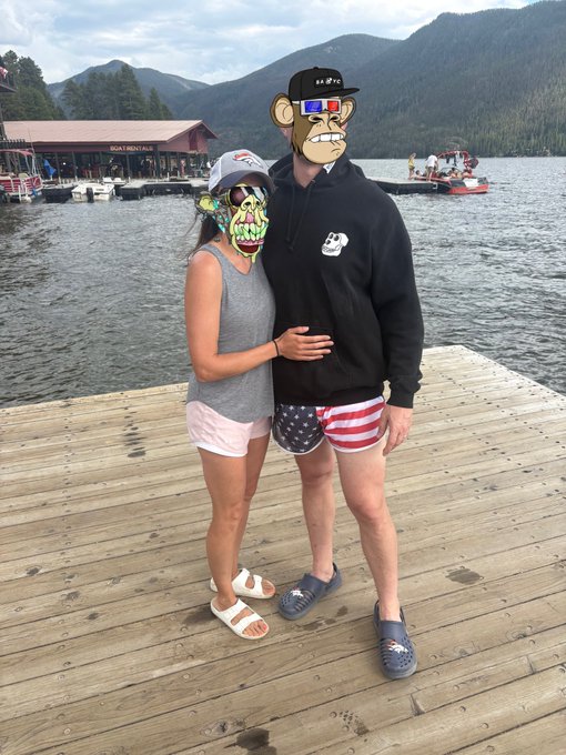 A couple poses on a wooden dock by a lake with boats and mountains in the background, their faces obscured by cartoon and skull avatars.