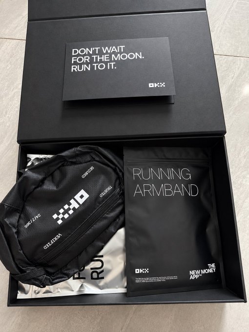 An open black gift box reveals a black running armband branded 'OKX' along with an OKX-branded drawstring bag. A card on top reads 'DON'T WAIT FOR THE MOON. RUN TO IT.'