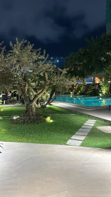 A serene nighttime view of a resort-style pool surrounded by lush greenery and stone pathways, with distant city lights twinkling under a cloudy sky. The tranquil setting contrasts with the post's urgent warning about the dangers of high leverage (x100), illustrating how quickly fortunes can vanish—just as a peaceful scene can be disrupted in an instant.