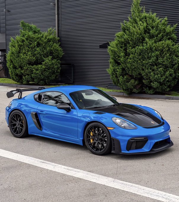 A Shark Blue 2024 Porsche 718 GT4 RS with Weissach Package sits on display, matching the post's details of a low-mileage, PPF-protected sports car with 8,900 miles and blue interior, listed at $185k with crypto accepted.