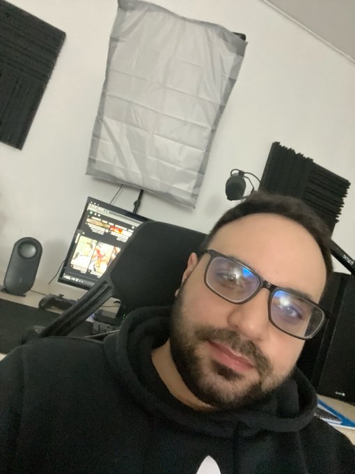 Selfie of a bearded man with glasses in a black hoodie, sitting in a room with acoustic panels, a computer monitor, and speakers in the background.