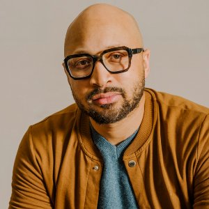 A portrait of a bald man with a beard wearing thick-rimmed black glasses and a brown jacket over a blue shirt.
