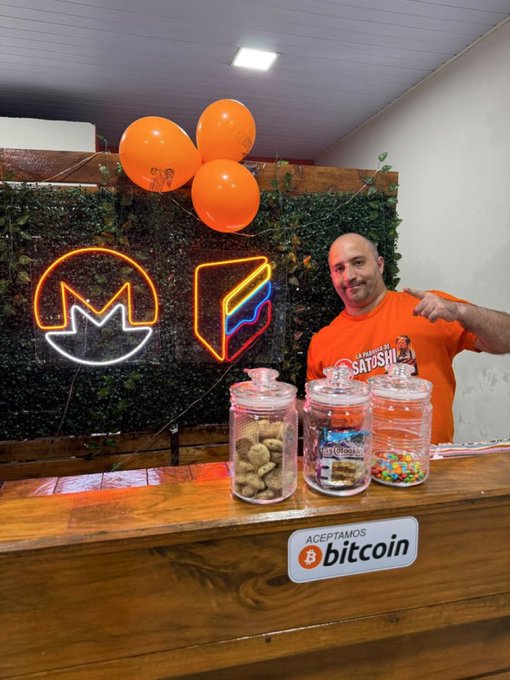 A man points to a display of sweets at an empanada stand in Paraguay that accepts Bitcoin and Monero, as indicated by the neon signs and "Aceptamos Bitcoin" sticker.
