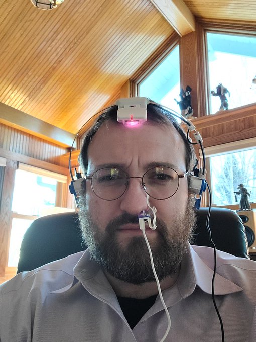 A bearded man with glasses wears a makeshift headset device with a glowing red sensor on his forehead and what appears to be a nasal probe, possibly jokingly referencing cryptocurrency founders with experimental brain-computer interfaces, while the accompanying post questions "Or cardano Founder?"