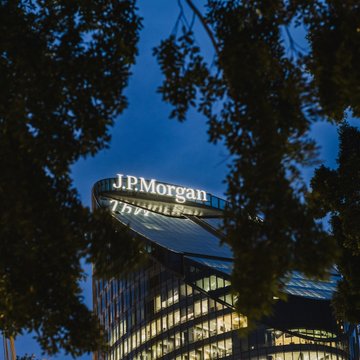A modern glass building displays the illuminated "J.P.Morgan" logo at its peak, framed by silhouetted tree branches against a twilight blue sky, visually representing the financial giant's institutional banking presence. The image ties to the news of JPMorgan expanding its JPM Coin deposit token to Ethereum for institutional clients.
