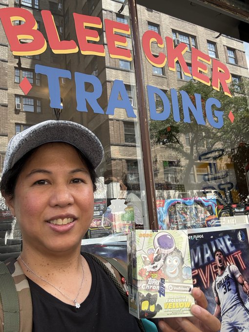 A woman wearing a grey cap stands in front of a Bleecker Trading store window displaying large red and blue lettering, holding Funko Pop! figures and a card.