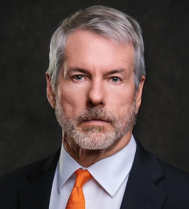 A portrait of a distinguished man with silver-gray hair and a beard, wearing a dark suit, light blue shirt, and bright orange tie, set against a dark backdrop. His confident expression aligns with the post's message about Michael Saylor, a prominent Bitcoin advocate, reinforcing the conviction behind his quote on Bitcoin clarity.