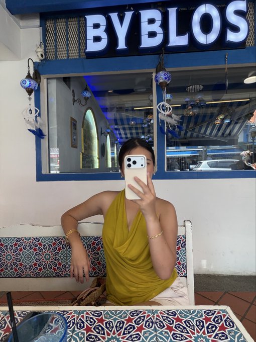 A woman takes a mirror selfie inside a restaurant called 'BYBLOS,' holding a phone with three cameras, wearing a yellow halter top and sitting at a table with patterned tile.
