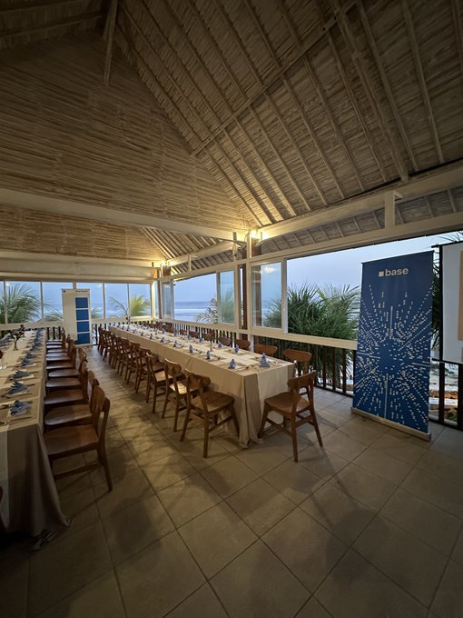 An empty event venue with a thatched roof, ocean views, and long banquet tables set for a "base" conference event.