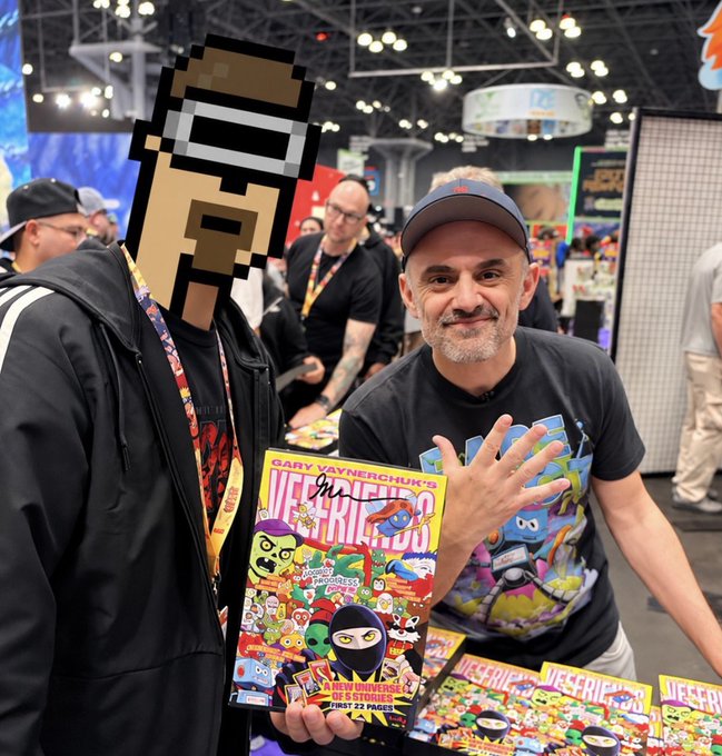 A man wearing a cap holds up a copy of "Gary Vaynerchuk's VeeFriends: A New Universe Part 2 Prints, First 22 Pages" comic book at what appears to be a convention. The image ties directly to the post's reference of "@veefriends," Gary Vaynerchuk's NFT and creative project, while the meme-style pixelated emoji face overlay captures the playful, grind-set energy of the caption.