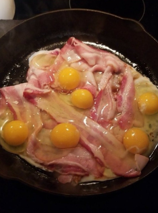 ```
A cast iron skillet contains strips of raw bacon topped with multiple cracked raw eggs, their bright orange yolks intact as the whites begin to cook.
```
