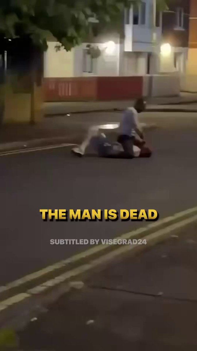 The image shows a man lying face down on the ground with his head above the street, possibly dead or injured. A person appears to be kneeling beside him, suggesting that they are attending to the situation. The surrounding area includes a sidewalk and a building in the background.