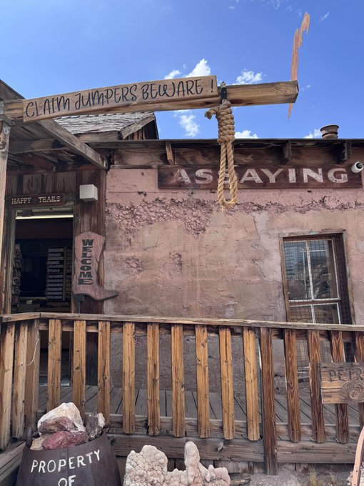 A rustic wooden building in a ghost town with weathered signs reading 'CLAIM JUMPERS BEWARE!', 'ASSAYING', 'HAPPY TRAILS', 'WELCOME', and 'PROPERTY OF' around a wooden porch with a hanging noose and rocks on display.