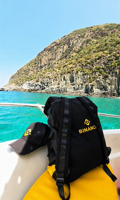 A black backpack and matching cap, both featuring the yellow Binance logo, are displayed on a boat with a dramatic rocky coastline and turquoise sea in the background, showcasing how the brand merchandise stands out beautifully against the sunlit seascape.