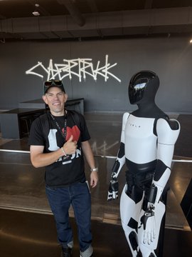 A man in a black "Cybertruck" t-shirt and baseball cap stands smiling next to a white Tesla Optimus humanoid robot inside a Tesla showroom, with a large "CYBERTRUCK" sign displayed on the wall behind them. The caption humorously suggests the human looks out of place without a cowboy hat, playfully implying that @grok could digitally add one to match the futuristic, sci-fi vibe of standing alongside the robot.