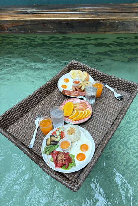 A floating breakfast tray holds plates of eggs, vegetables, fruit, and rice, along with glasses of orange juice, served in a swimming pool.