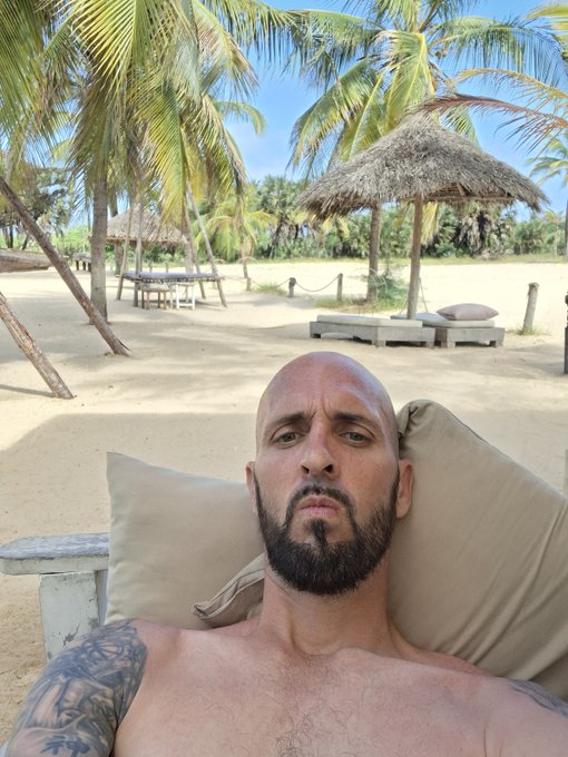 A shirtless bald man with a beard and tattoos relaxes on a lounge chair at a tropical beach with palm trees and a thatched-roof hut.