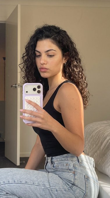 A young woman with curly dark hair takes a mirror selfie in a black tank top and light blue jeans, holding a phone with a purple beaded case.
