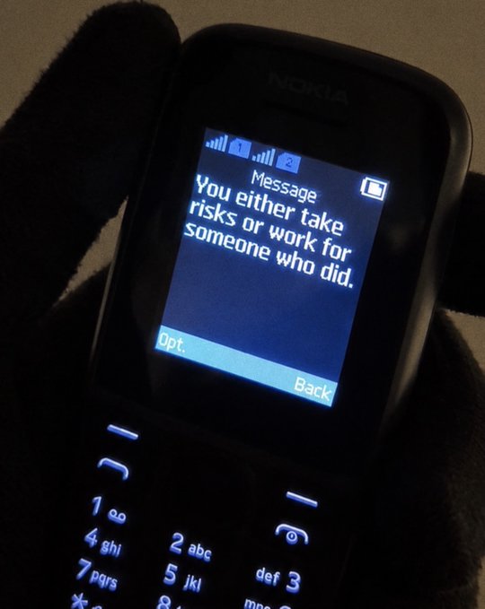 A gloved hand holds a Nokia feature phone with an illuminated blue screen displaying a message: 'You either take risks or work for someone who did.'