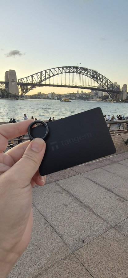 A hand holds a black Tangem card up against the iconic Sydney Harbour Bridge at sunset, with the city skyline and harbor visible in the background. The image ties into a Tangem social media contest inviting users to share real photos of their cards or rings in meaningful locations.