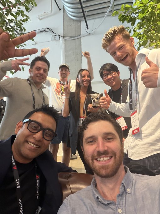 A group selfie of six people posing indoors with one person holding a small dog, with some wearing lanyards indicating an event or conference.