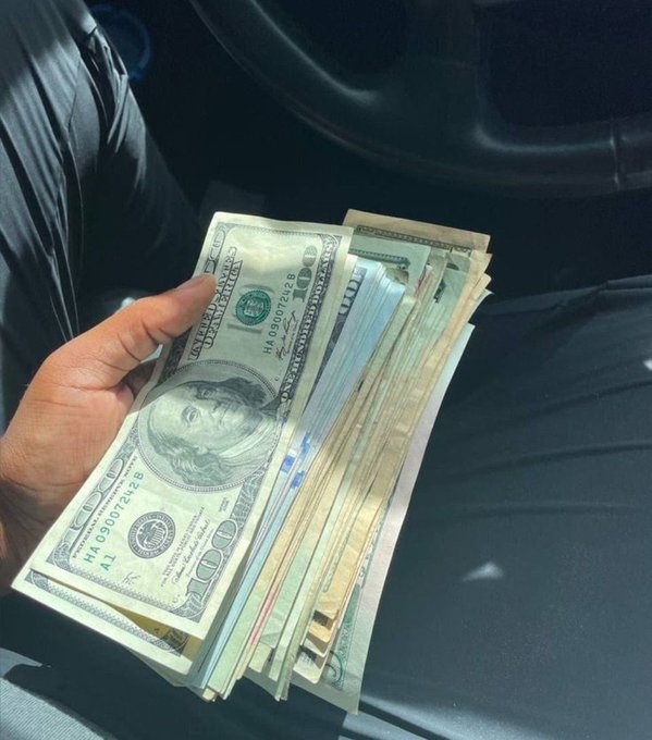 A hand holds a fanned-out stack of U.S. $100 bills inside a car, with the visible bill showing serial number "HA 09900742 B," illustrating the post's hopeful sentiment about a profitable day.