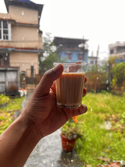 A hand holds up a glass of milky chai against a backdrop of traditional buildings and a rainy garden, embracing a peaceful morning ritual that pairs perfectly with a relaxed "Gm legends" greeting to the community.