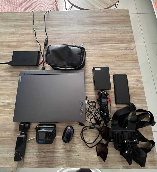 A travel/mobile work setup laid out on a wooden table includes a Lenovo Legion 5 laptop, external hard drives, GoPro camera, DJI Osmo Pocket, Zoom recorder, sunglasses, a body harness/chest mount, and a power bank with cables.