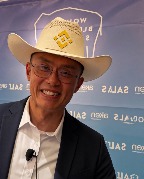 A smiling man wearing glasses, a white shirt, dark blazer, and a cream-colored cowboy hat with a yellow logo, standing in front of a backdrop that reads 'TOKEN 2049 SALT LAKE' and 'OMAKTAU'.