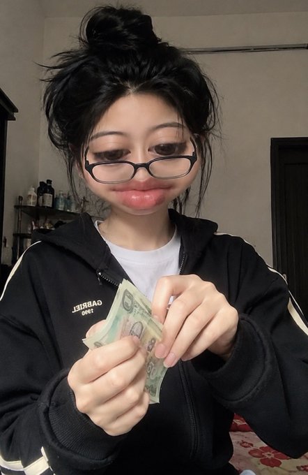 A young woman with dark hair in a messy bun, wearing glasses and a black zip-up hoodie over a white shirt, holding a stack of Thai baht banknotes.