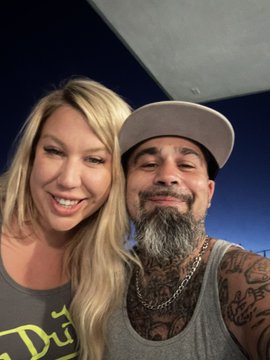 A selfie of a smiling blonde woman and a bearded man wearing a baseball cap, both in tank tops, against a dark blue background.