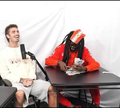 A man in a red outfit and orange beanie sits at a table counting a thick stack of cash while another man laughs beside him, capturing a moment of casual wealth display that mirrors the post's boastful tone about profiting off a token and investing the gains into "Ansem."