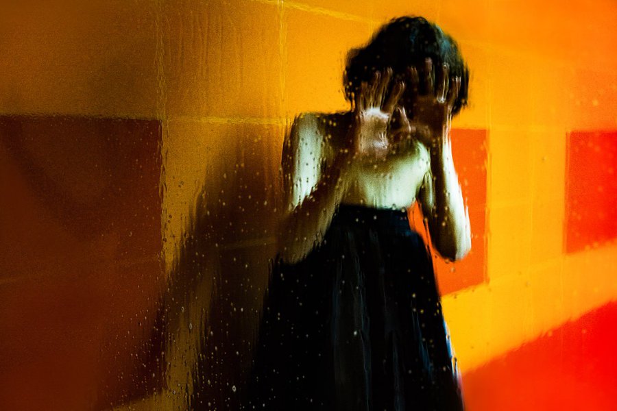 A stylized, atmospheric image of a figure with hands covering their face, viewed through a wet, rain-spattered glass with warm orange and yellow lighting.