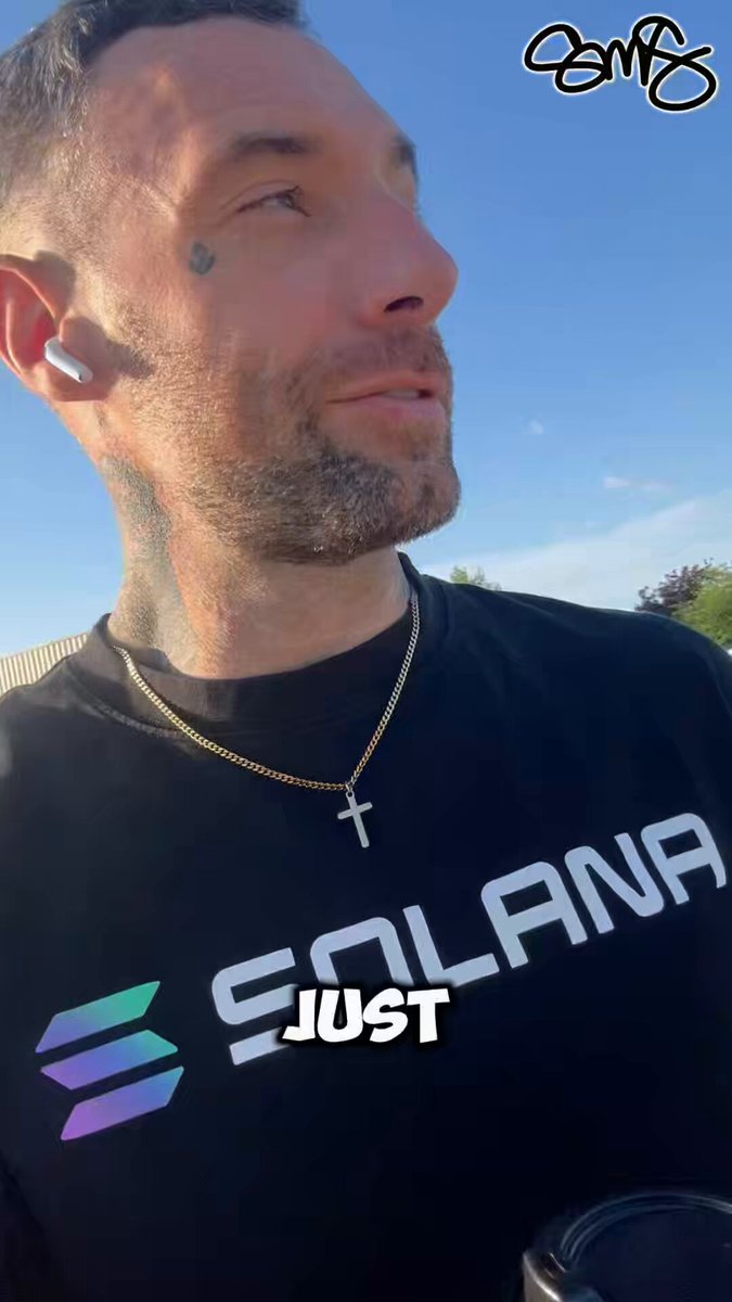 The image features a man wearing a black shirt with the word "Solana" written across it, along with a cross necklace. He has a beard and is looking up at something above his head. The caption of the image reads "@solana".