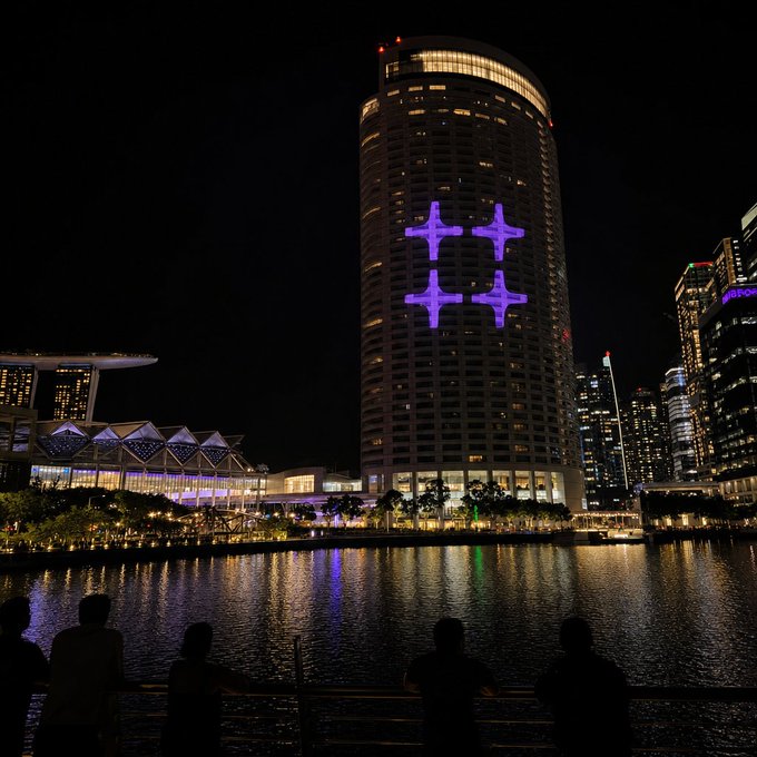 A nighttime cityscape featuring a tall cylindrical skyscraper illuminated with purple drone lights forming hashtag-like symbols, with silhouettes of people watching from across a body of water.