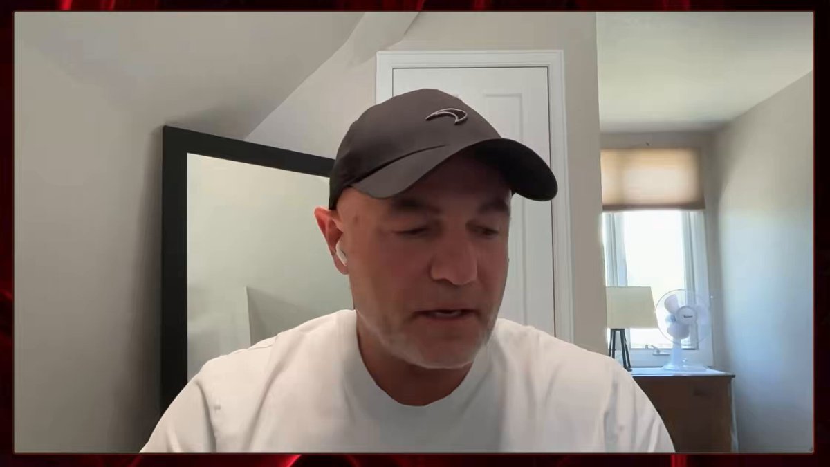 The image shows a man wearing a black baseball cap sitting at a desk with his face partially visible behind him. The background features a window that allows natural light into the room, creating an atmosphere of comfort and relaxation. There are also two chairs present in the scene, one near the center of the image and another towards the right side.