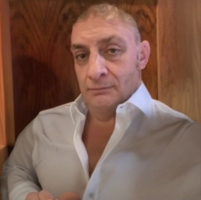 A selfie-style photo of a middle-aged man with short hair wearing an open-collared white shirt, seated in front of wood-paneled walls.