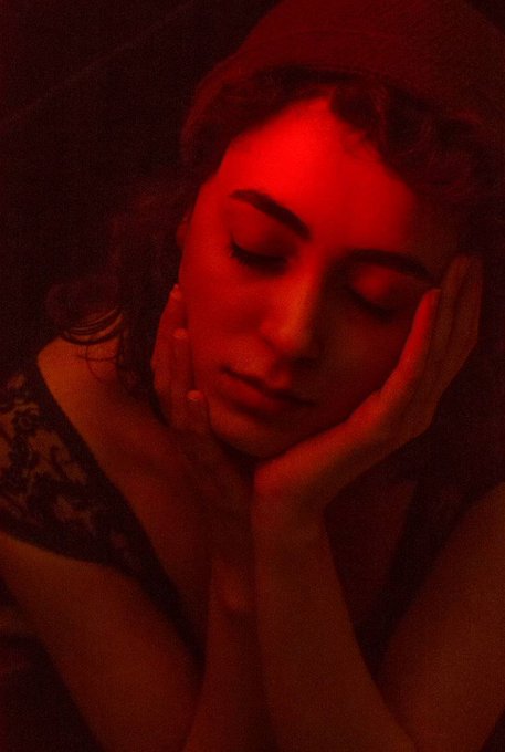 An artistic red-tinted photograph of a woman with her eyes closed, resting her face against her hands in a serene, contemplative pose.
