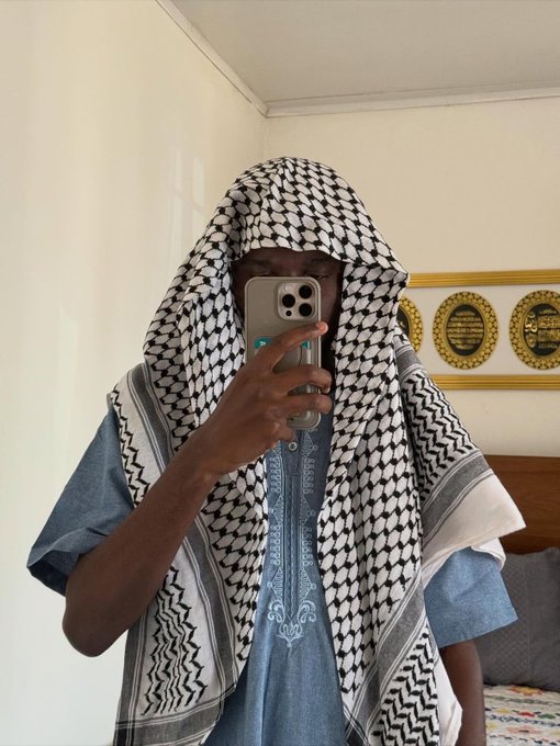 A person takes a mirror selfie while wearing a blue traditional outfit paired with a black and white patterned keffiyeh draped over their head.