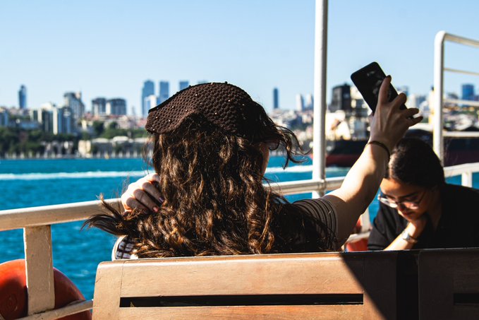 A young woman with long curly brown hair and a black beanie takes a selfie on a boat crossing blue waters, with a city skyline visible in the background. Another passenger sits nearby as the urban landscape stretches across the horizon.