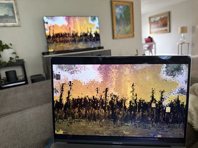 A MacBook Pro displaying a glitchy, pixelated landscape artwork on a living room couch, with a TV in the background showing the same or similar image.