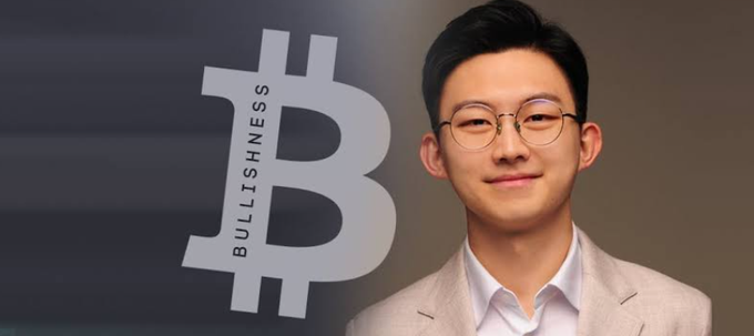 A young man with short black hair and glasses, wearing a beige blazer and white shirt, stands in front of a backdrop featuring a large Bitcoin "₿" symbol with the word "BULLISH" displayed vertically, representing a more tempered outlook on $BTC's bull cycle returns (x3-x5) compared to previous cycles.