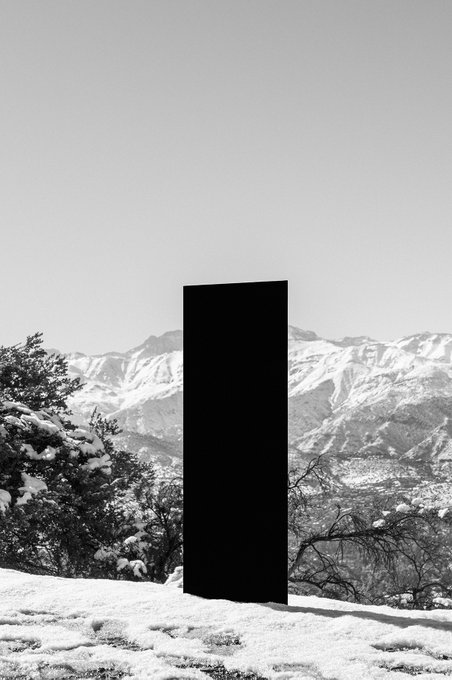 A tall, sleek black monolith-like portal stands stark against a snowy mountain landscape, embodying the "Snow Portals 001" series by @TransientLabs. The dark rectangular form contrasts dramatically with the pale sky and snow-dusted terrain, evoking a surreal sense of an otherworldly gateway in nature.